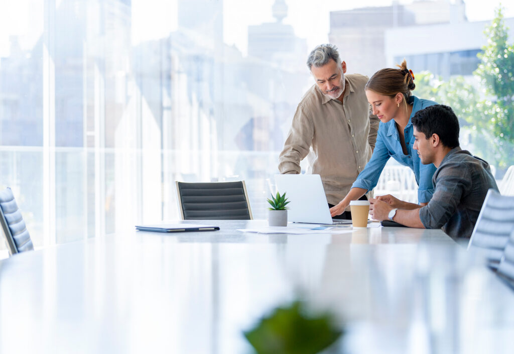 Three colleagues collaborate around a laptop in a bright, airy office conference room